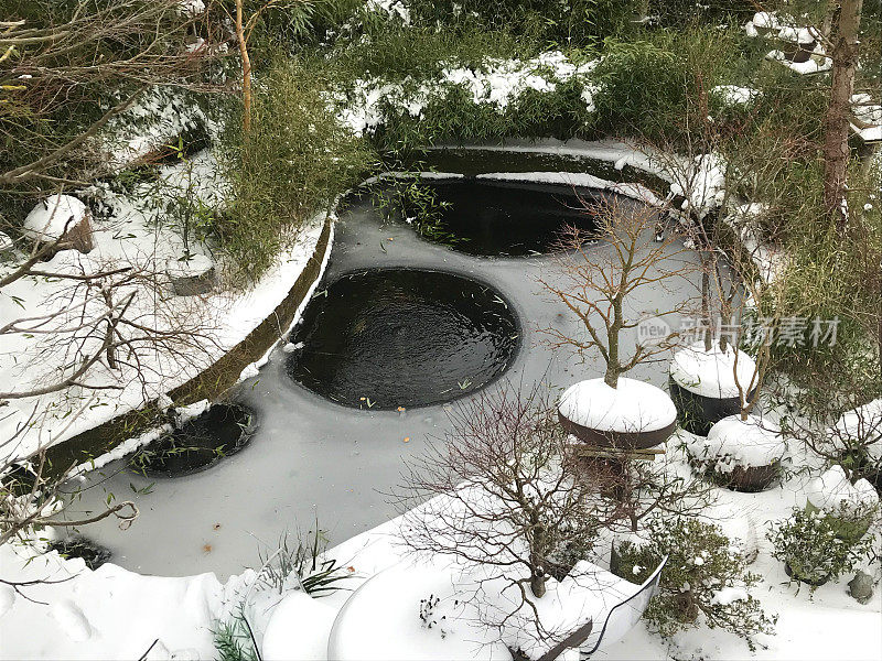 大冰封的锦鲤池在冬天的花园被冰雪覆盖后，大雪/暴风雪，金鱼池泵与曝气气泡阻止水冻结的东方日本园林盆景树和禅宗灯笼